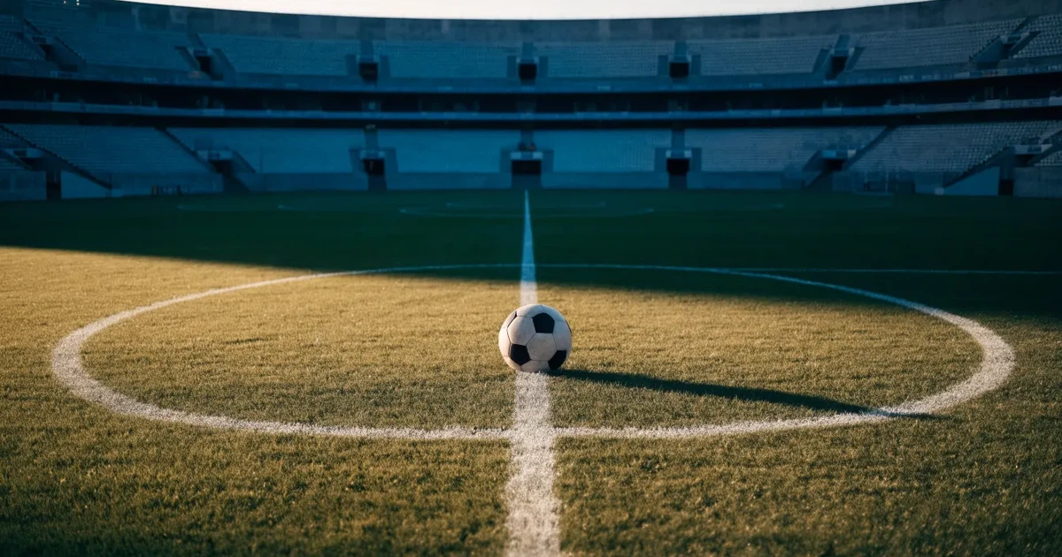 Ein Fußball liegt auf dem Anstoßpunkt eines leeren Rasenplatzes, im Hintergrund eine große Stadiontribüne im Abendlicht.
