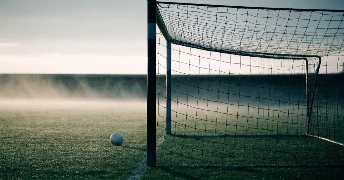 Ein Fußballtor mit gespanntem Netz steht auf einem leeren Rasenplatz, davor ein einzelner Ball, Morgendunst liegt über dem Feld.