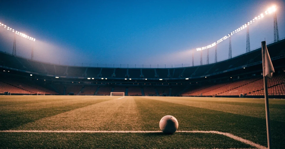 Blick durch ein Stadiontor auf einen grünen Rasen im Abendlicht, ein Ball liegt an der Eckfahne, die Ränge sind leer.