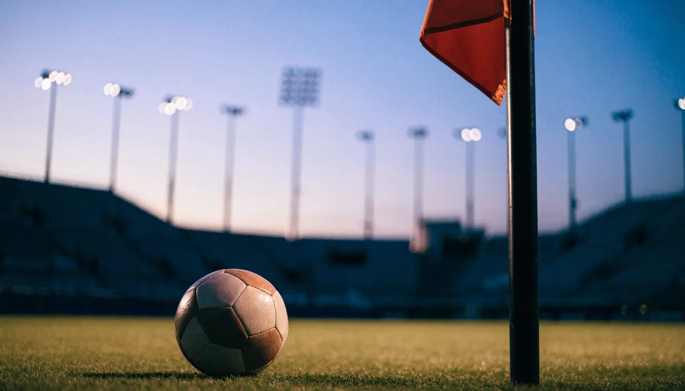 Ein Fußball liegt neben einem Eckfahnen-Pfosten auf einem Rasenplatz, im Hintergrund ein leeres Stadion in der Dämmerung.