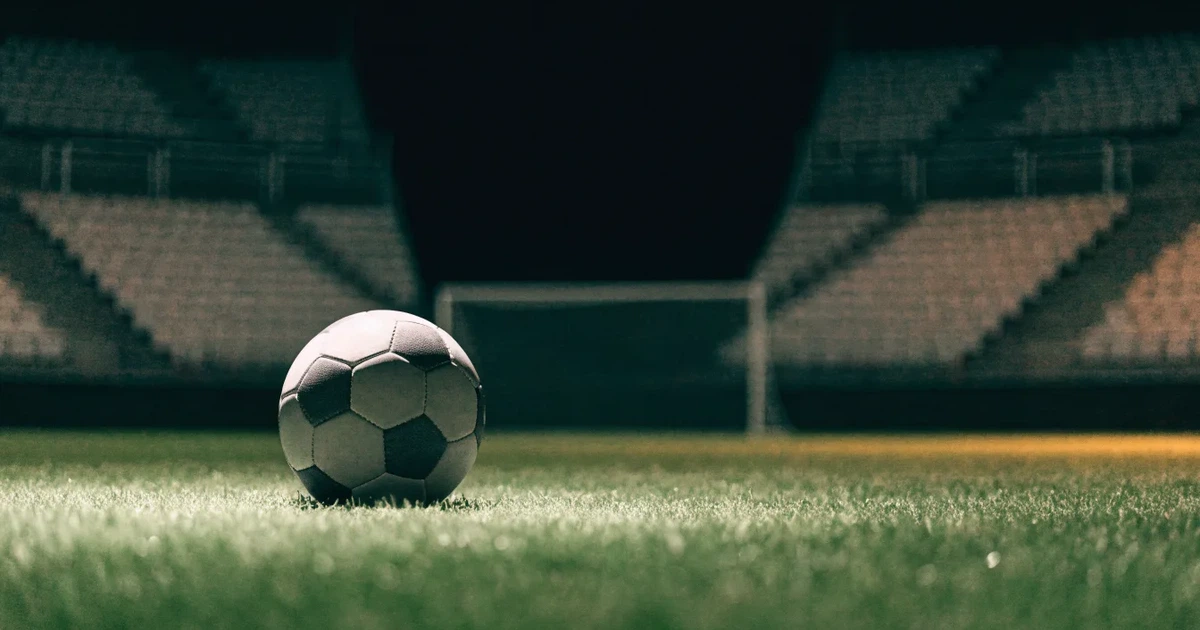 Ein Fußball liegt auf dem Rasen eines leeren Stadions bei Flutlicht, im Hintergrund unscharfe Tribünen ohne erkennbare Beschriftung.