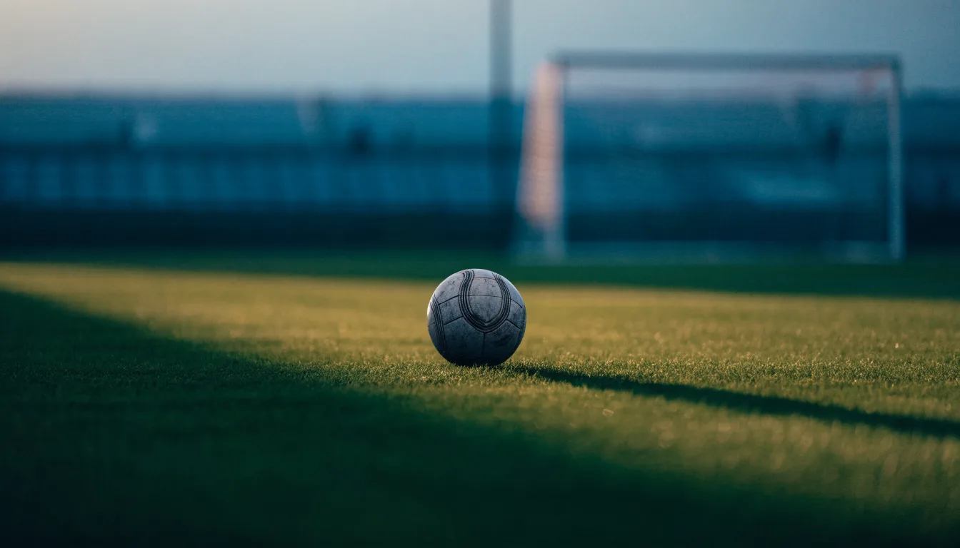 Ein Fußball liegt im Strafraum eines leeren Platzes, dahinter die Netze des Tores im Abendlicht.