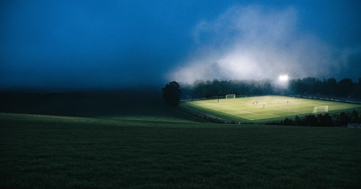 Ein Stadion unter Flutlicht, aus großer Entfernung über eine dunkle Wiese fotografiert, am Himmel zieht Nebel auf.