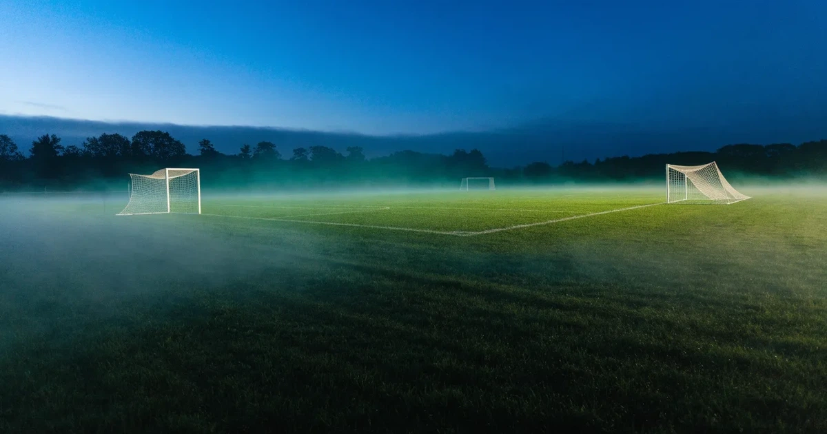 Ein Stadion unter Flutlicht, aus großer Entfernung über eine dunkle Wiese fotografiert, am Himmel zieht Nebel auf.