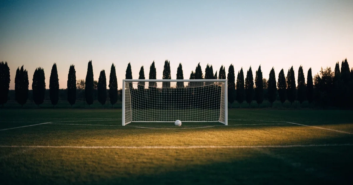 Ein Fußballtor mit Netz auf einem leeren Platz im Abendlicht, der Rasen frisch gemäht, im Hintergrund Zypressen.