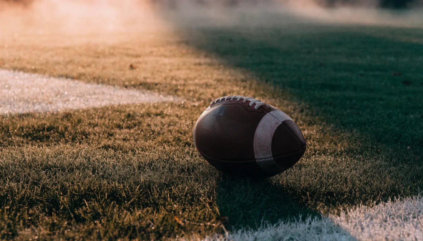Ein ovaler Lederball mit heller Schnürung liegt auf dem Rasen vor der Endzone, Tau auf dem Gras im ersten Morgenlicht.