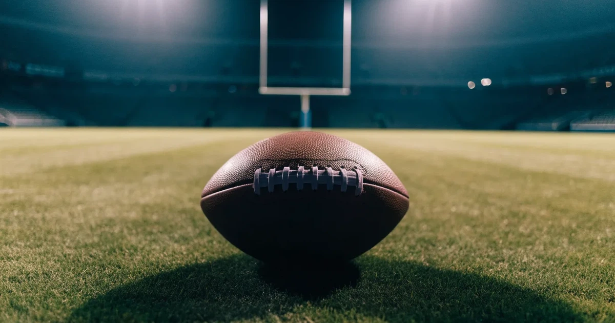 Ein American Football liegt auf dem Rasen eines leeren Stadions unter Flutlicht, die Torstangen ragen im Hintergrund auf.