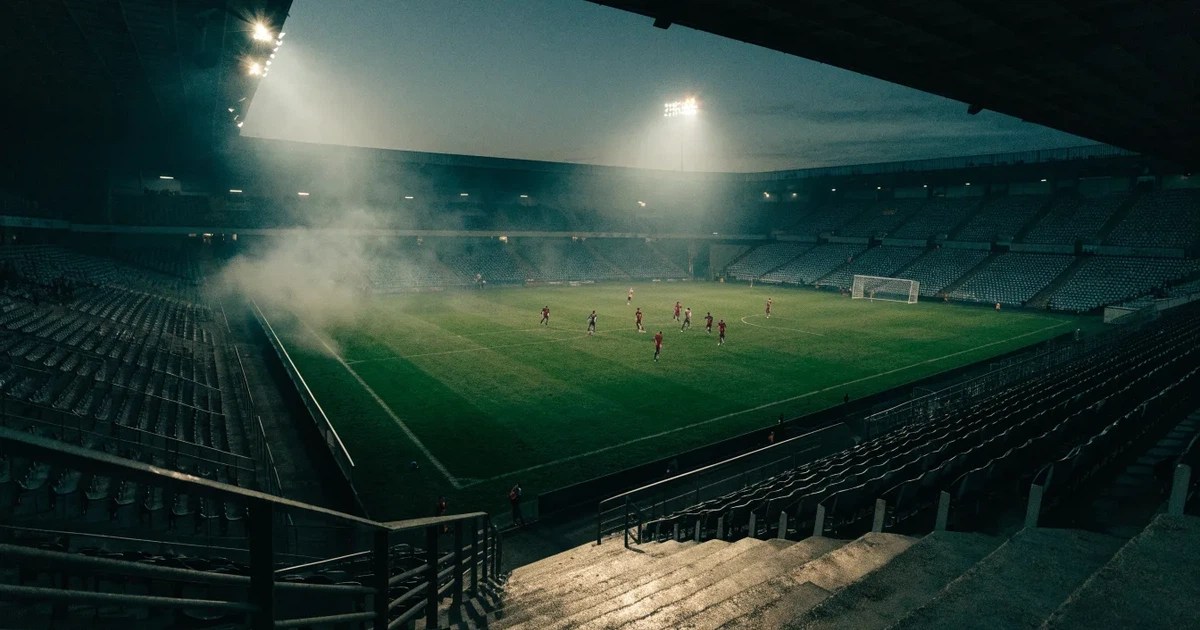 Ein leeres Stadion unter Flutlicht aus großer Entfernung gesehen, Dunst liegt über dem Rasen, die Tribünen sind nur als Lichtpunkte erkennbar.