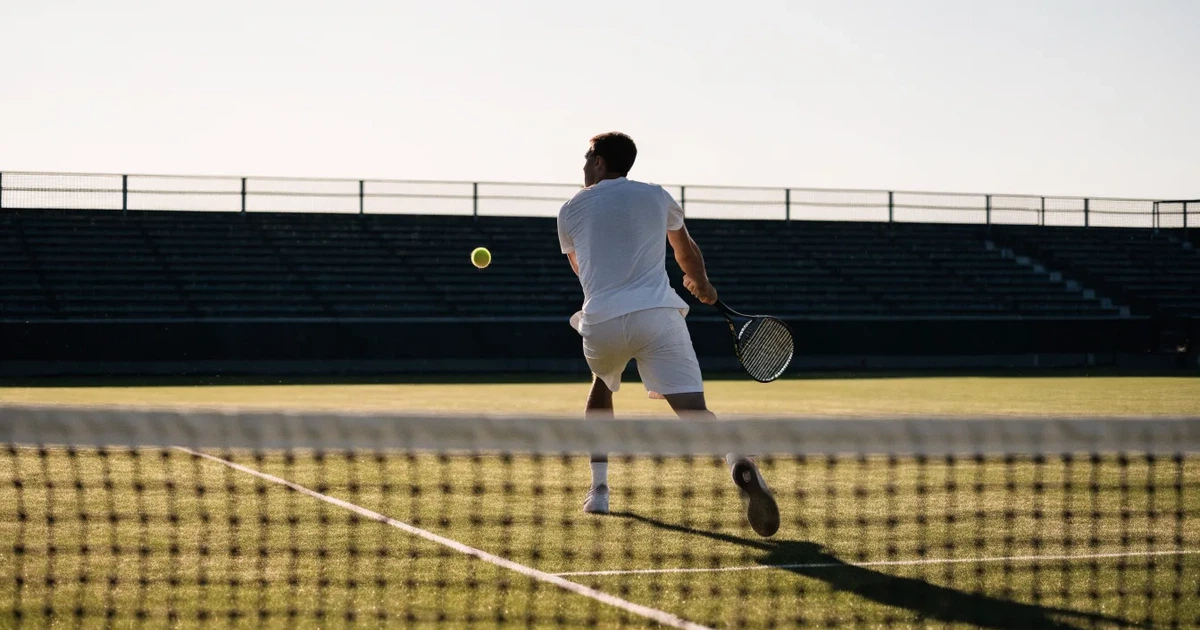 Ein Tennisplatz mit Rasenbelag im Morgenlicht, Netz und weiße Linien, im Hintergrund eine ruhige Tribüne.